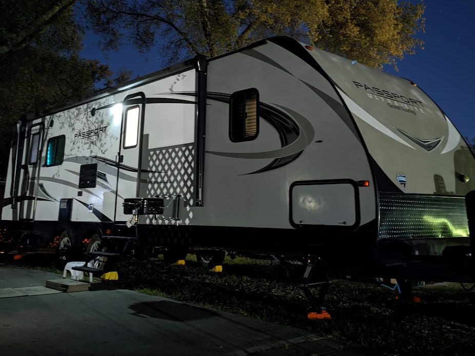 A travel trailer lit up at night, parked beneath tall trees with stars in the sky above it